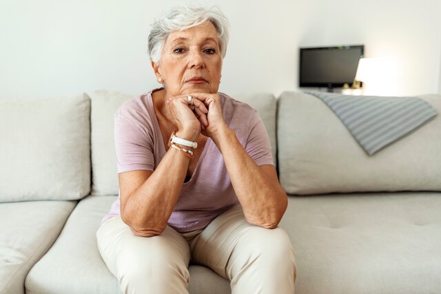 Eine ältere Dame mit weißen kurzen Haaren sitzt auf einem Sofa und sieht ernst in die Kamera.